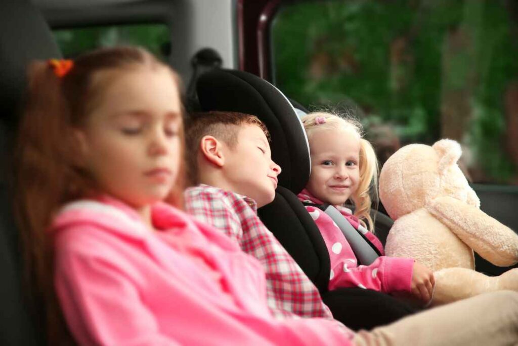 Three kids in the back seat, one holding a teddy bear.