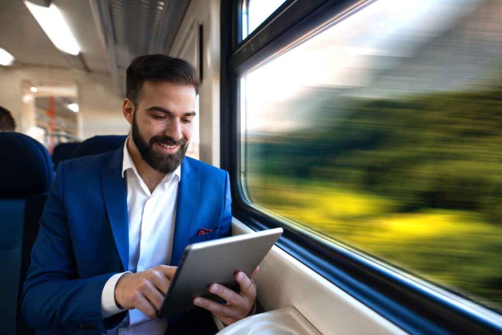 man in suit sitting by train window looking at a tablet and smiling