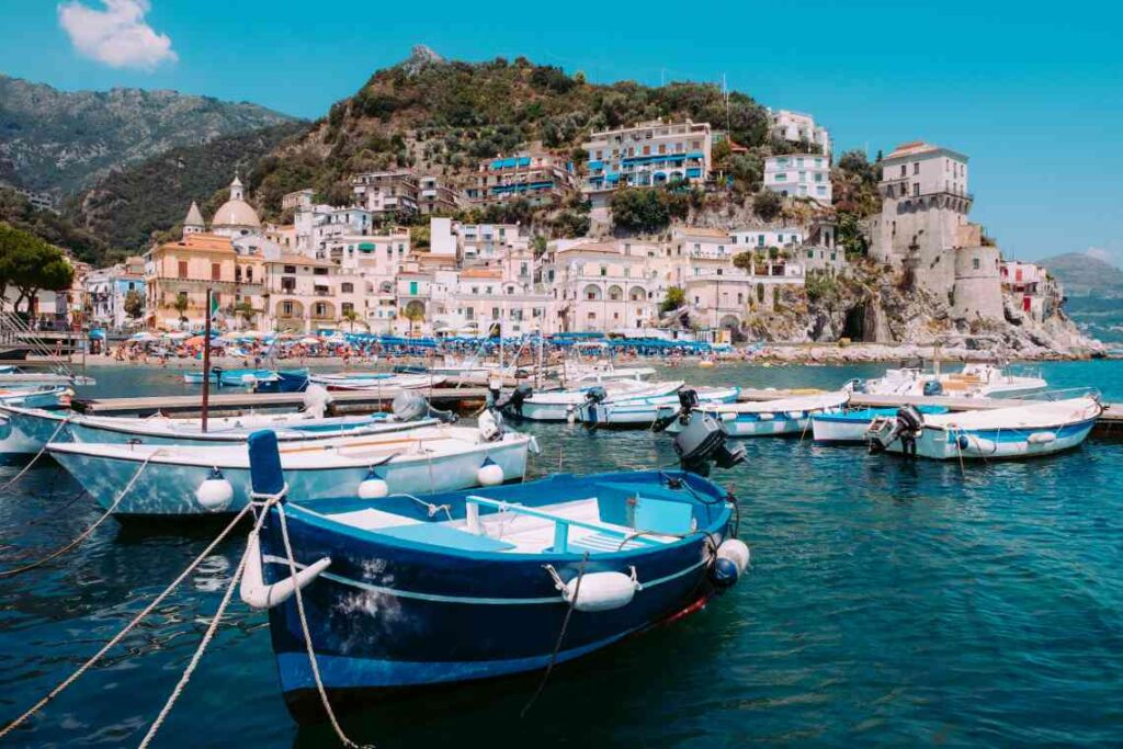 blue and white fishing boats on the water in front of a beach and small Italian village