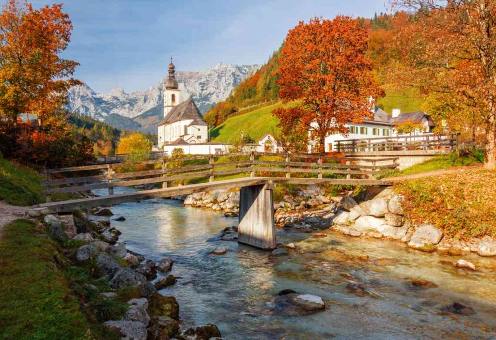 wooden bridge over a small river in Ramsau Germany with white church and mountains in the background