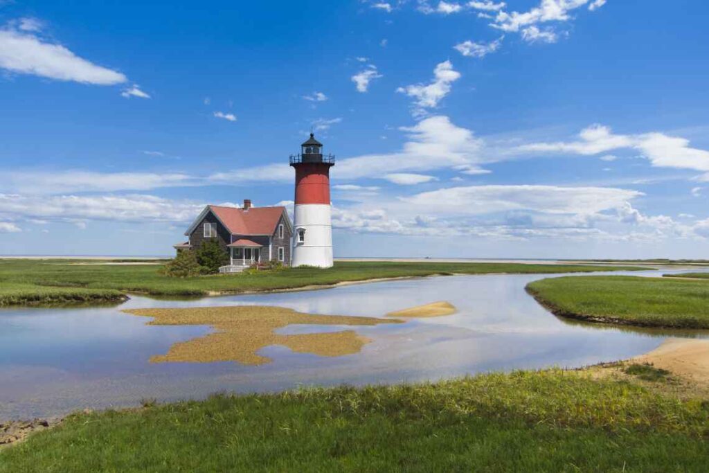 Traditional gray and white house next to a white and red lighthouse on a bog with green grass.