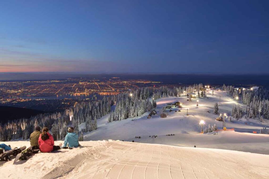 three people with snowboards sitting atop grouse mountain ski hill at sunrise