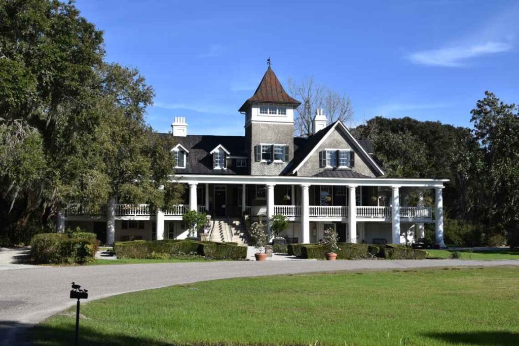 large gray and white house with columns and wraparound porch with driveway in front and sculpted bushes