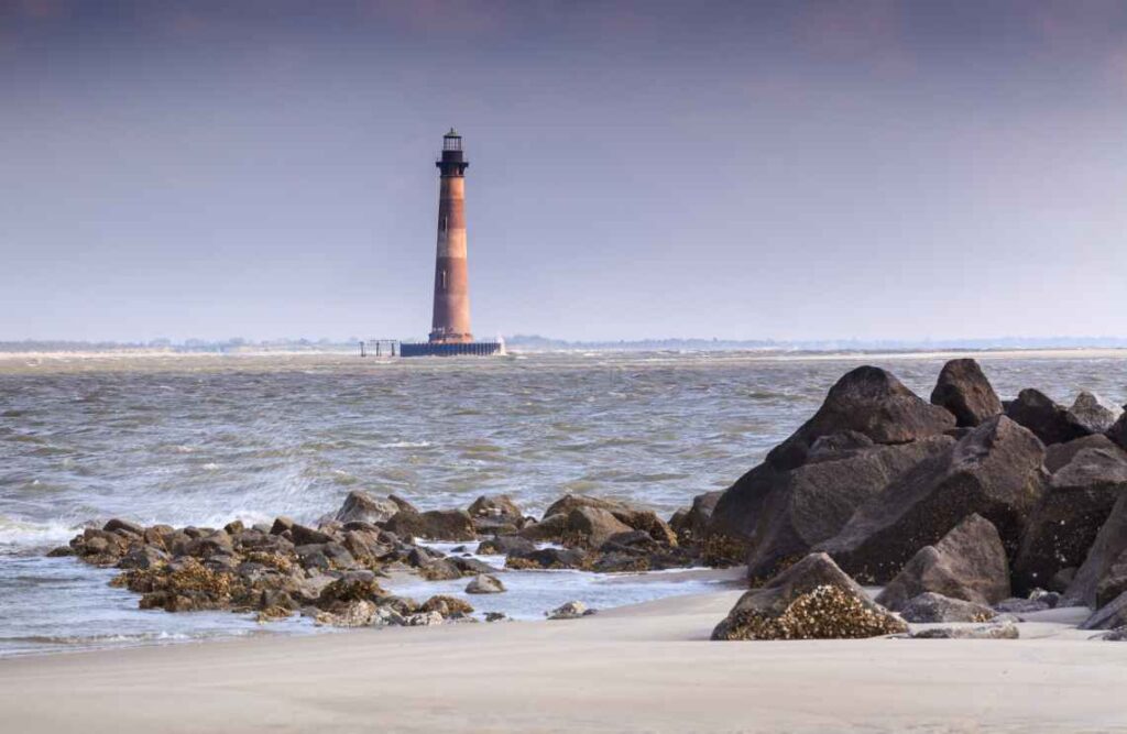 faded white and red lighthouse sitting off the coast with a beach and large black rocks in front