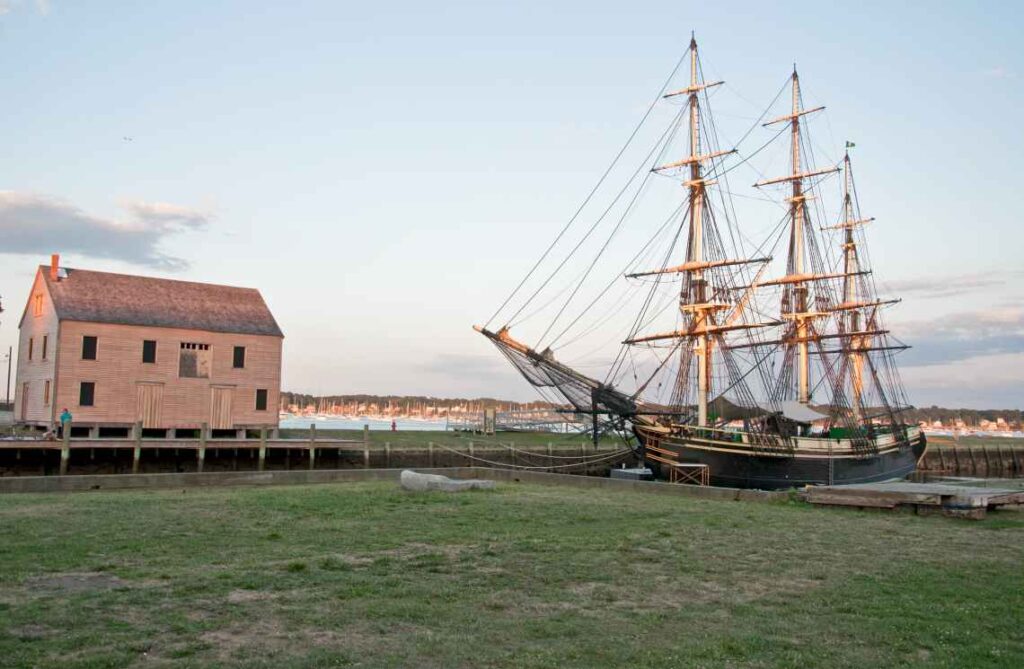 old ship with 3 masts docked in a harbor next to an old wooden building