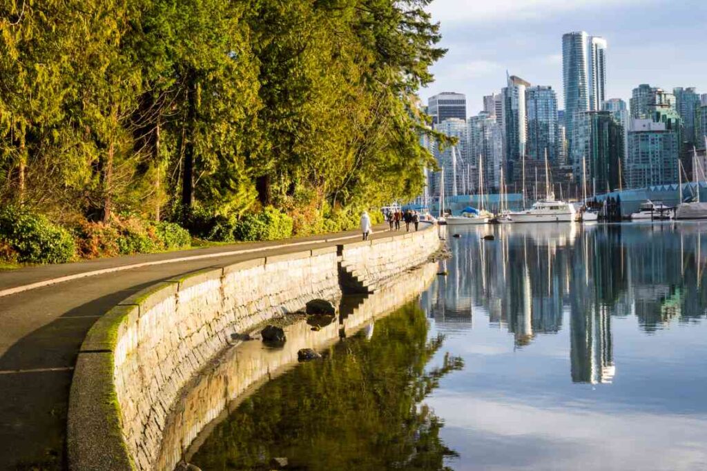 green trees and paved pathway along the water with boats and vancouver skyline in the background