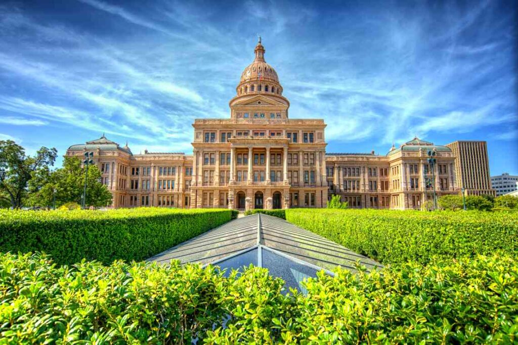 tan stone texas state capitol building with green hedges in front and blue sky with white clouds behind