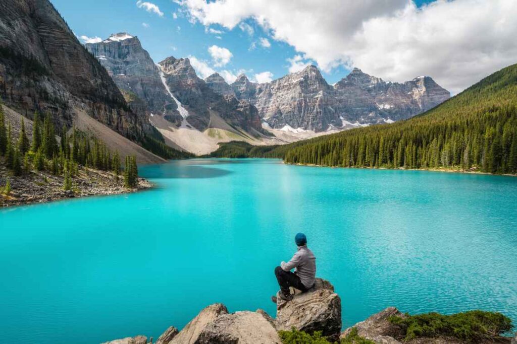 Hiker sitting on a rock in front of turquoise Moraine Lake with mountain peaks in the background and green forest