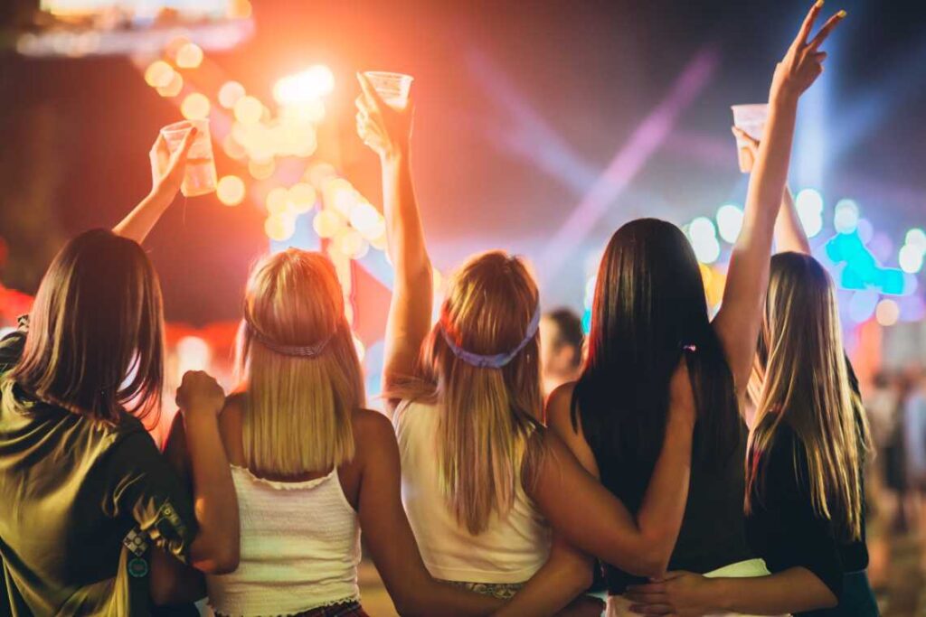 five women with their backs to the camera at a concert and their hands in the air holding drinks
