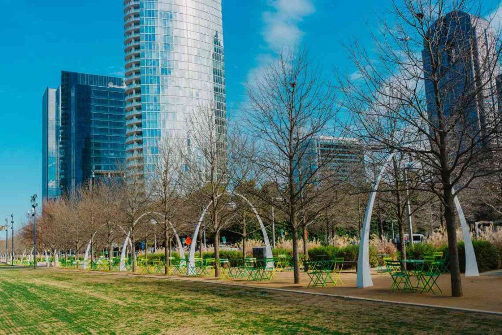 park with white arches and green tables and chairs with trees without leaves in front of skyscrapers in dallas