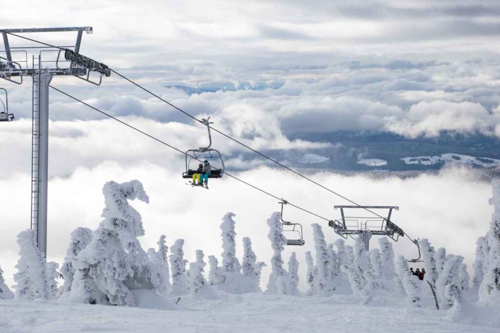 Skiers on an open ski lift above the clouds and snow covered pine trees