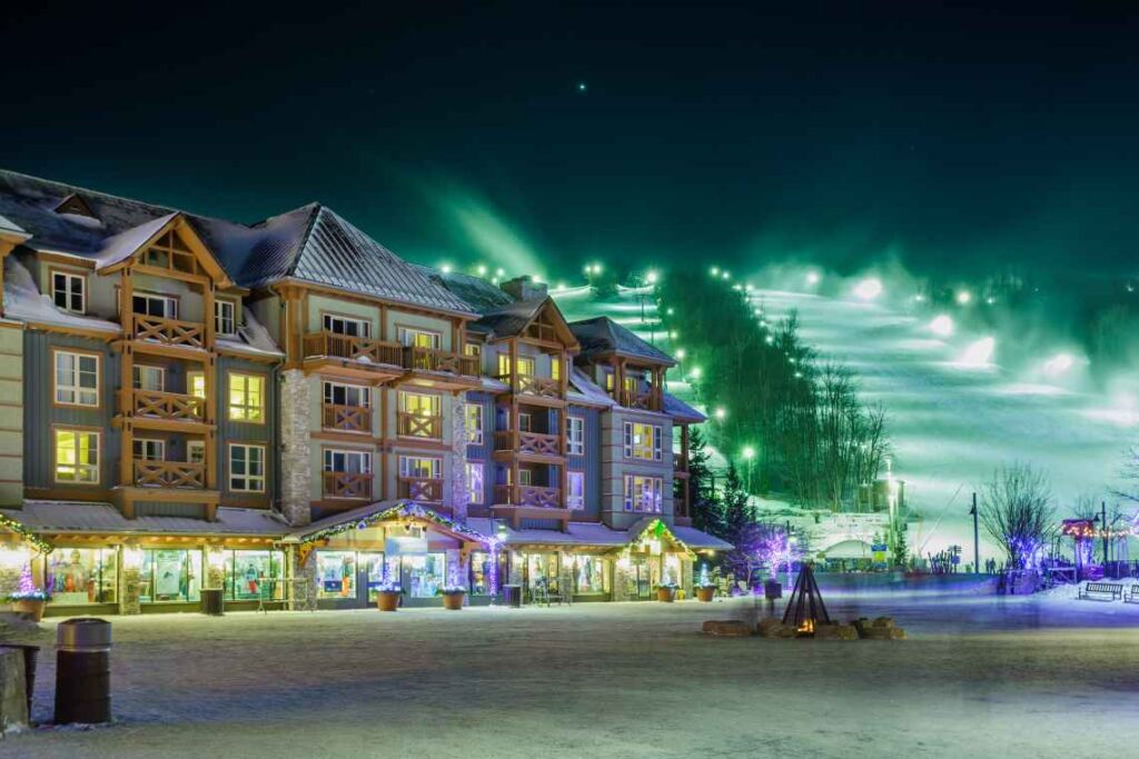 lighted ski runs rising behind a ski resort building with snow and shops on the ground floor
