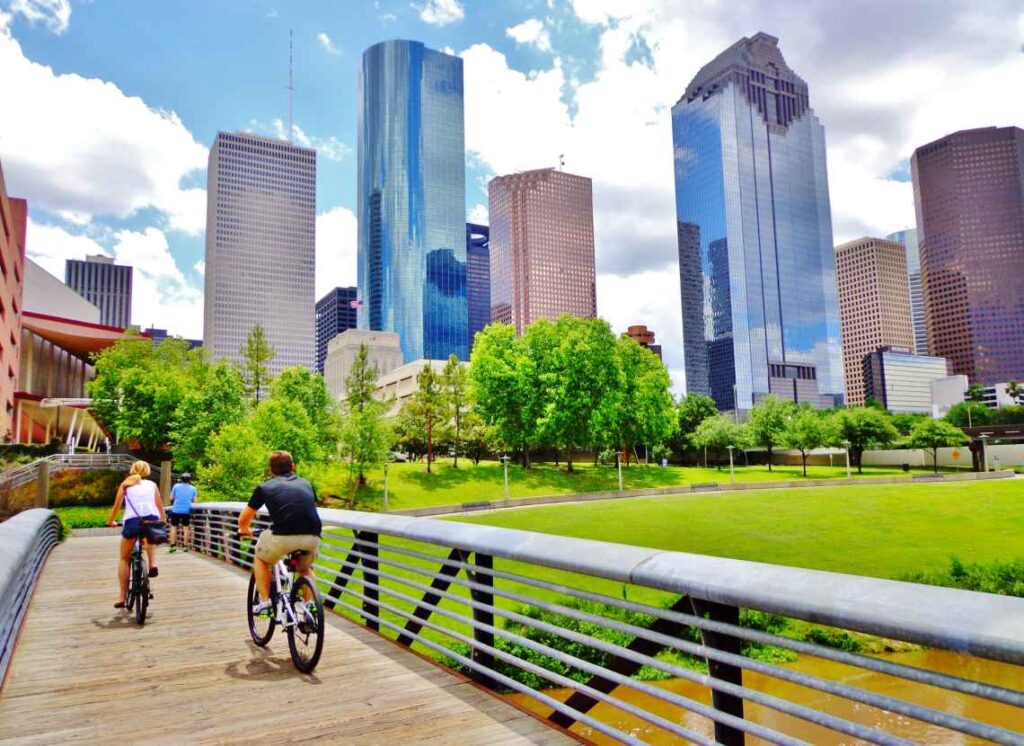 people riding across a wood and metal bridge over brown water with green grass and skyscrapers in the background