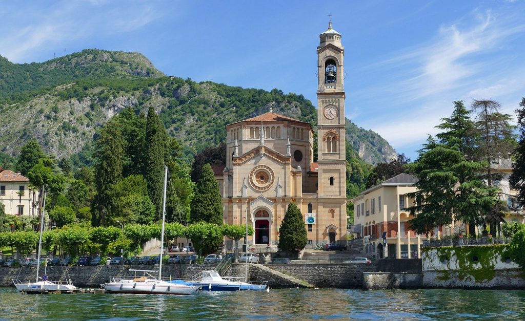 stone church on the shore of lake como with sailboats on the water in front and green mountains behind