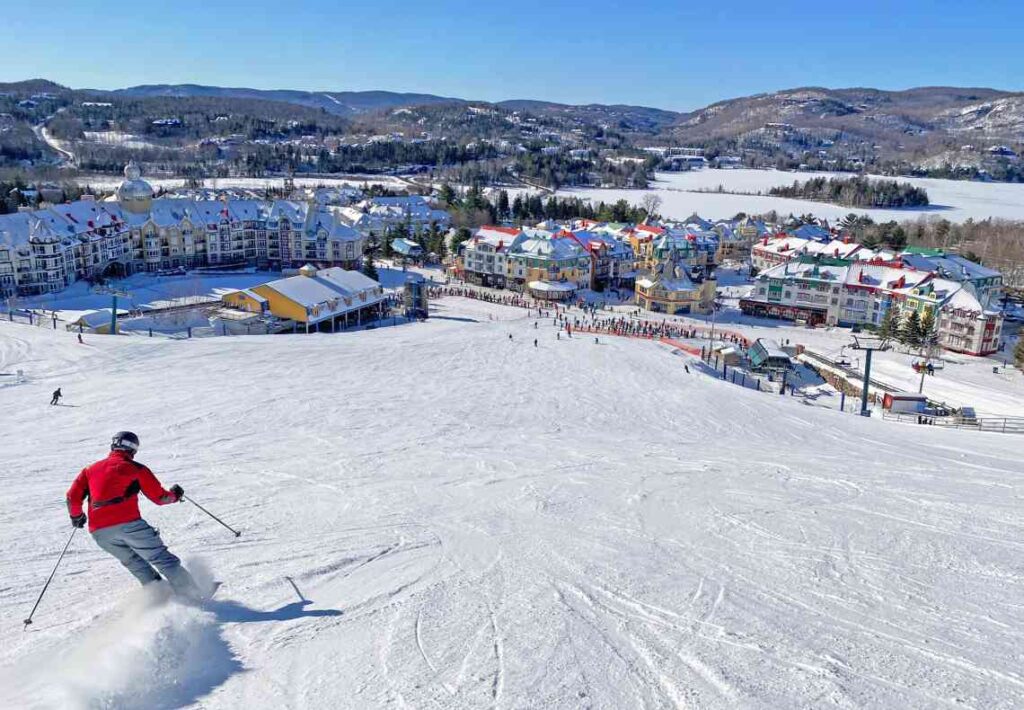 Person in red jacket and gray pants skiing down a hill toward a resort village