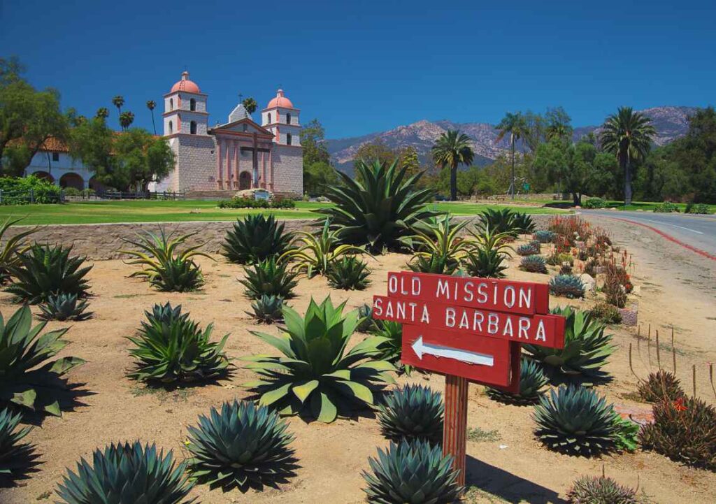 sign saying old mission santa barbara pointing toward the pink and white mission with cactus