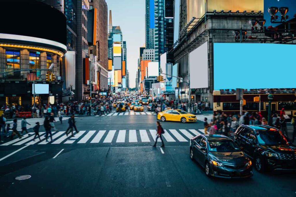 Bilden visar Times Square med ett övergångsställe i centrum. Människor korsar gatan och de flesta bilarna väntar vid ett trafikljus. 