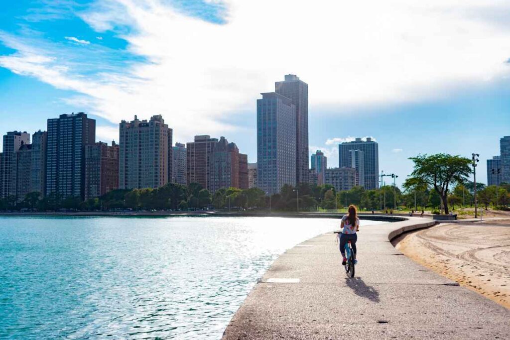 Uma mulher andando de bicicleta em um caminho de concreto ao longo do lago michigan com arranha-céus