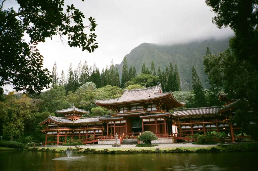 Japanese style red and white temple with a pond in front, with trees and green mountains rising in the background