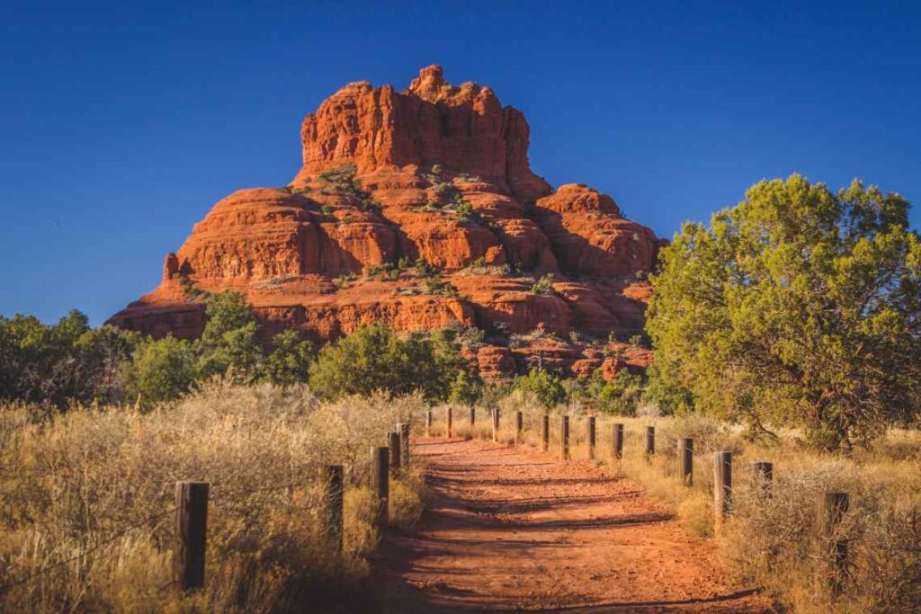 red rock structure of bell rock with a red dirt path leading to it and trees and tall grass around