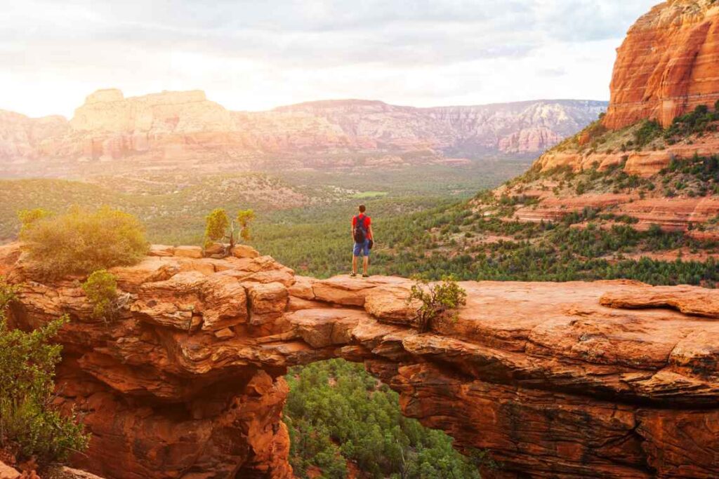 man in red shirt and blue shorts wearing a black backpack standing on devil's bridge looking over the red rock landscape