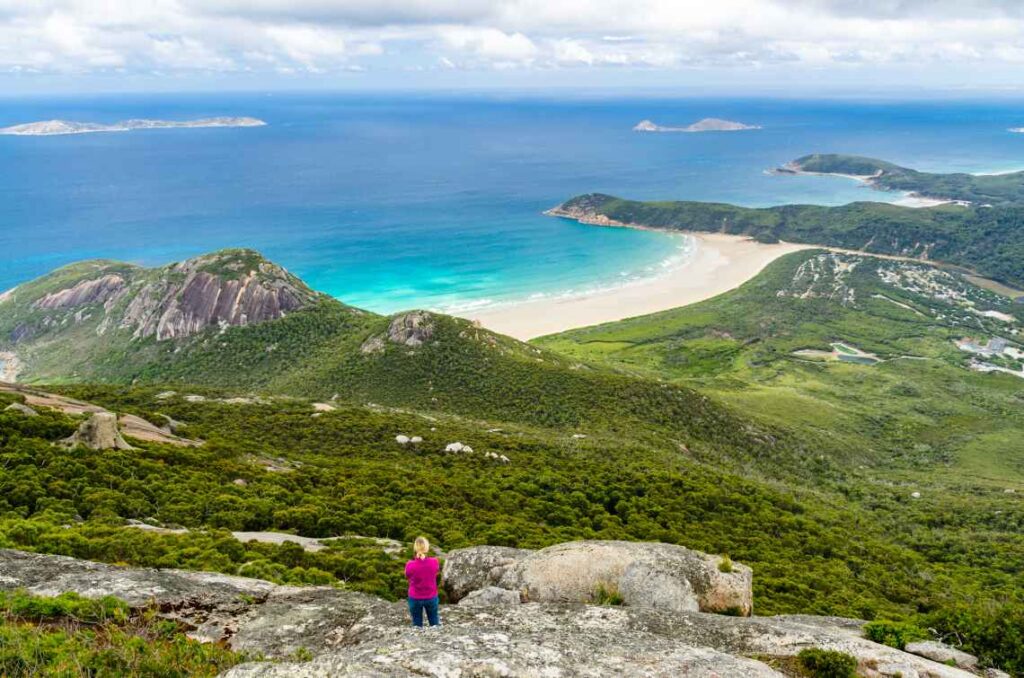 blond woman in purple jacket and blue pants standing on a rock looking over the ocean in wilsons promontory national park