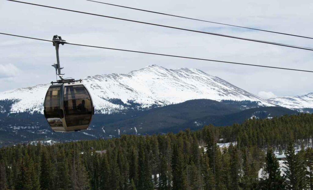 enclosed glass gondola going over snowy terrain with pine trees, and snow-covered mountains in the background