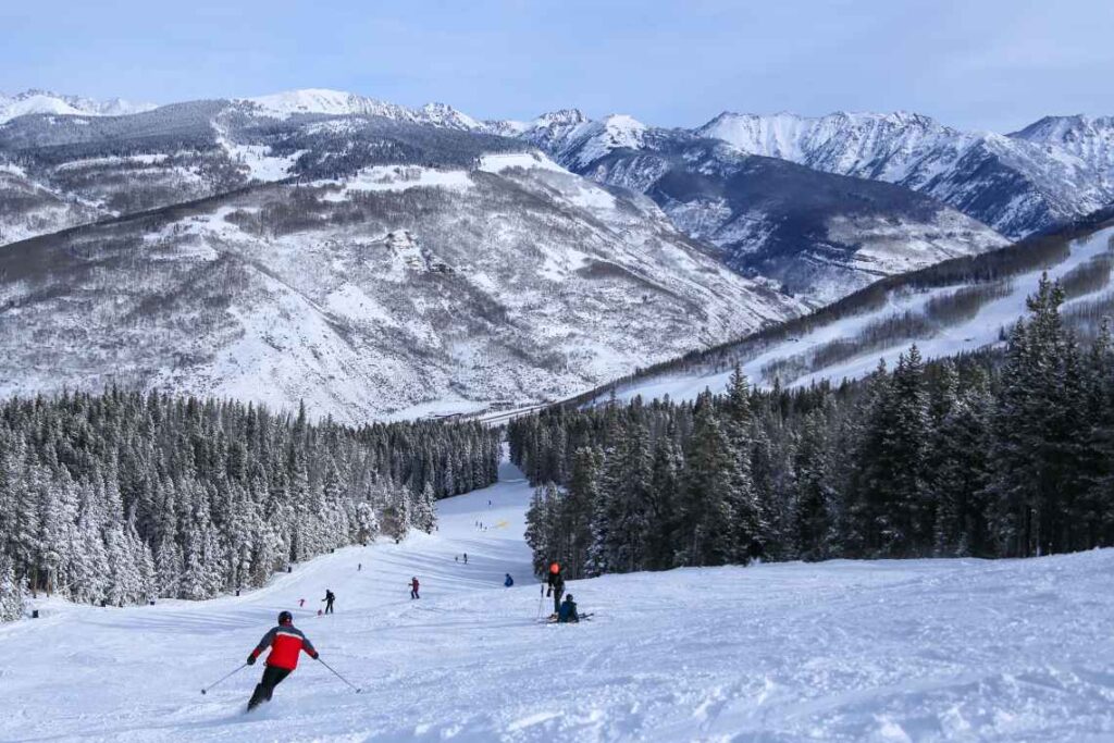skiers going down a snow-covered trail at the vail ski resort in colorado with trees and mountains in the background