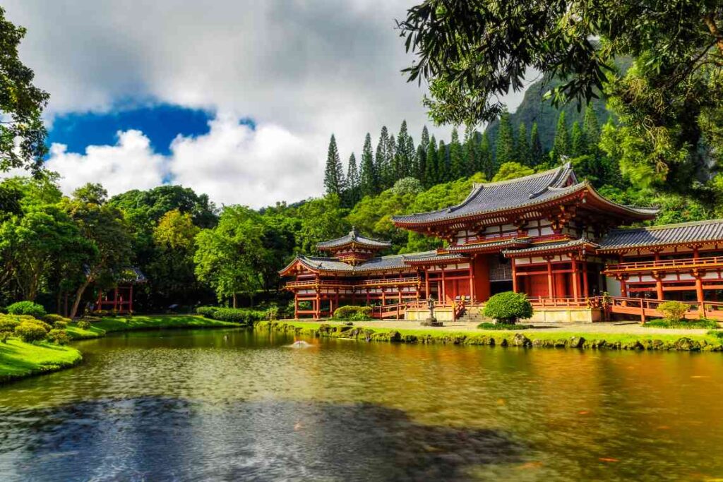 red and white japanese-style temple in front of a pond and with lush greenery, heavy clouds and mountains behind