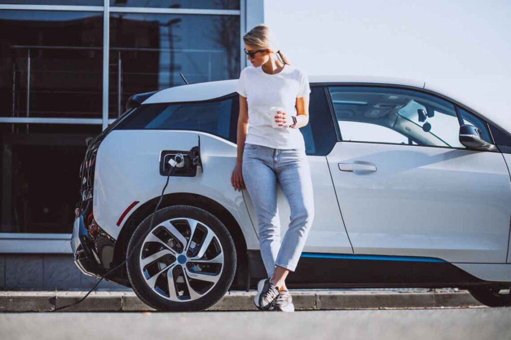 mulher com óculos de sol segurando uma xícara de café em pé na frente de um carro elétrico BMW branco que está conectado e carregando