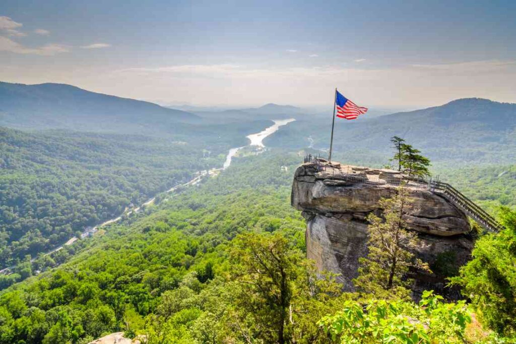 chimney rock jutting out of the green forest in chimney rock state park with an american flag flying on top