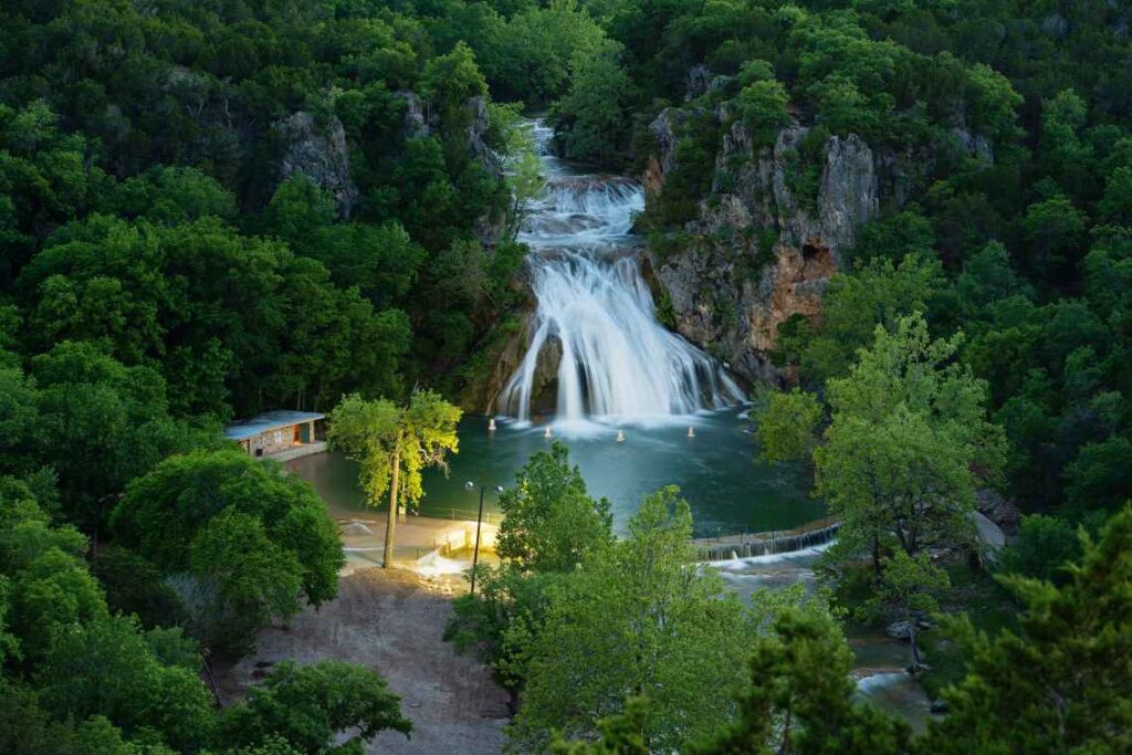 waterfalls flowing down rocks into a blue pool in the middle of a forest with rocky mountains