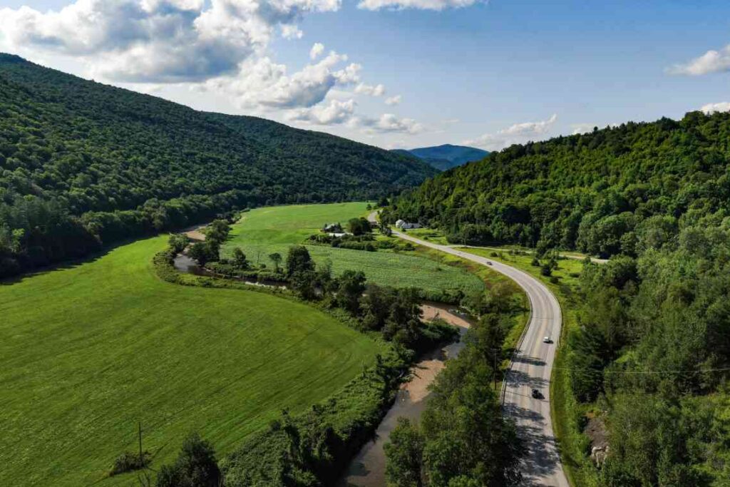 highway running through green grass and tree-covered mountains on a sunny day in vermont