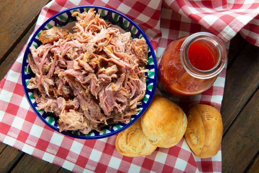 bowl of pulled pork with white bread rolls and a bottle of red vinegar-based sauce sitting on a red and white checked cloth