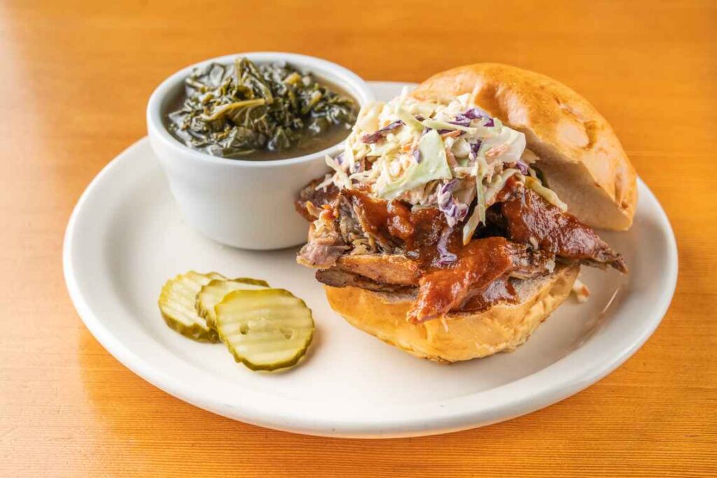 pulled pork barbecue sandwich with coleslaw on a white plate with a side of greens