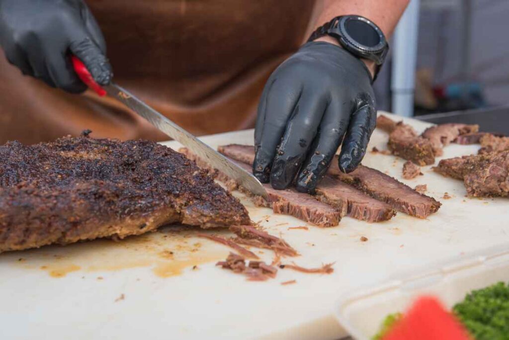 hands wearing black latex gloves slicing brisket with a knife