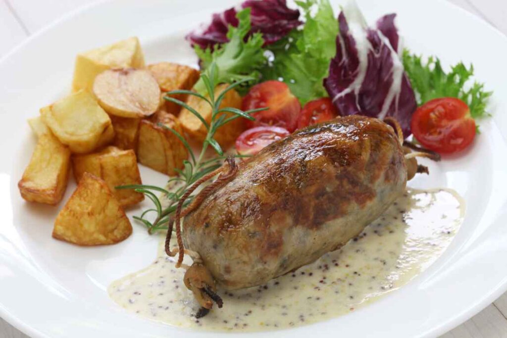 close up of lyon france style sausage sitting on creamy sauce with roasted potato pieces and green salad on a white plate