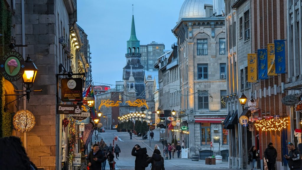 A lively pedestrian street in Old Montreal lined with restaurants, cafes, and decorative lights at dusk.