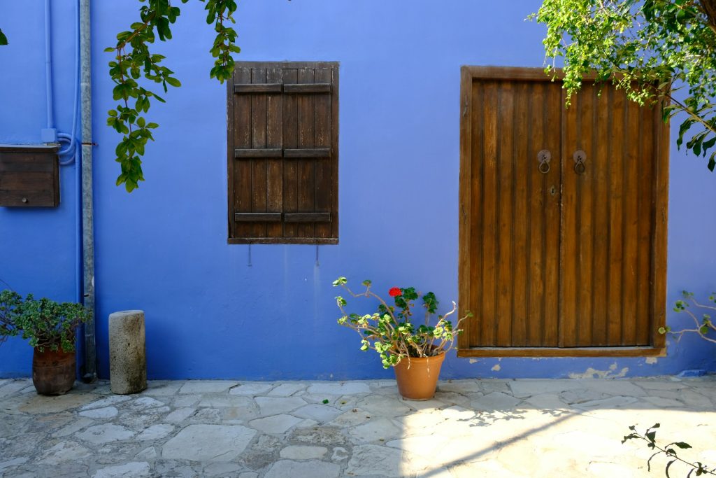 Bright blue wall with wooden door, shutters and a potted flower on a stone patio.