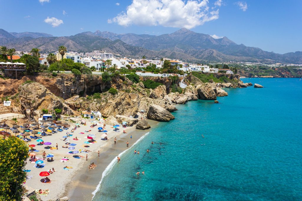Busy beach in Nerja with turquoise water, colorful umbrellas and rocky cliffs under a blue sky.