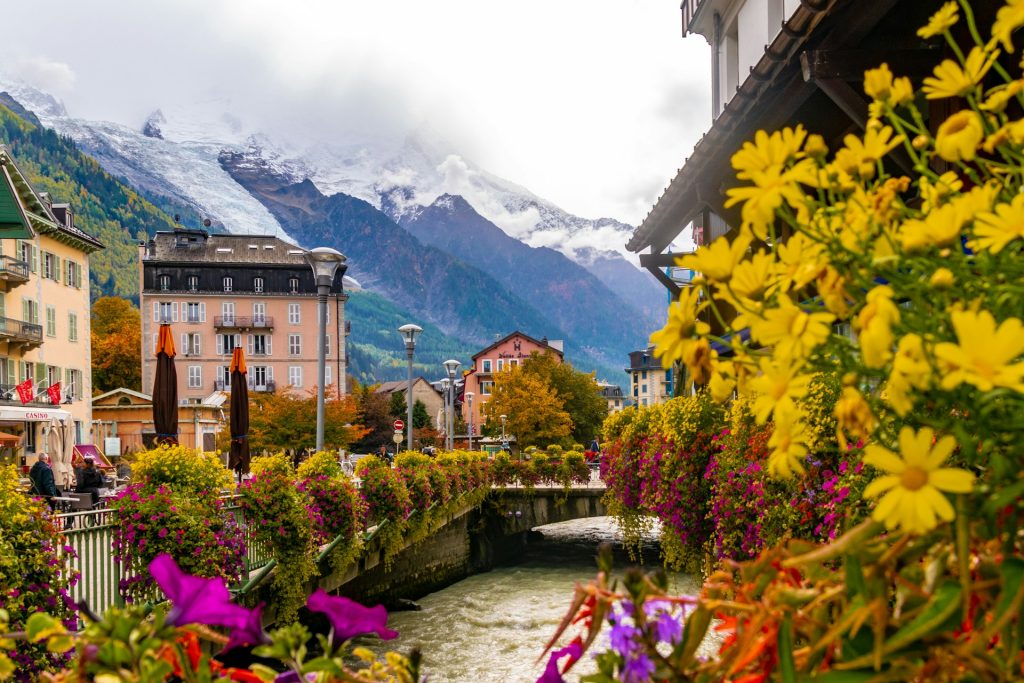 Flower-lined river in Chamonix with colorful buildings and snow-capped Mont Blanc massif in the background.