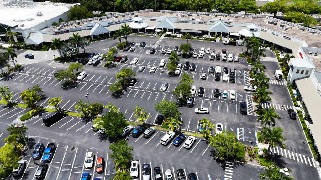 Aerial view of a large outdoor parking lot with palm trees and a row of shops in the background.