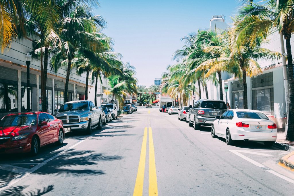 Cars parked on a sunny palm-lined street with shops and blue sky.