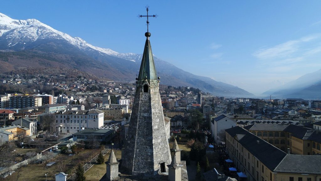 Stone church tower with cross in Aosta, surrounded by the city and snow-capped Alpine mountains.