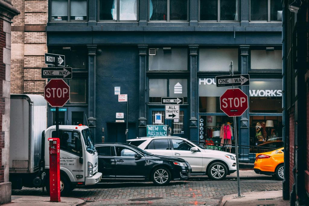 Busy city street corner with stop signs, one-way signs and parked cars outside a row of shops.