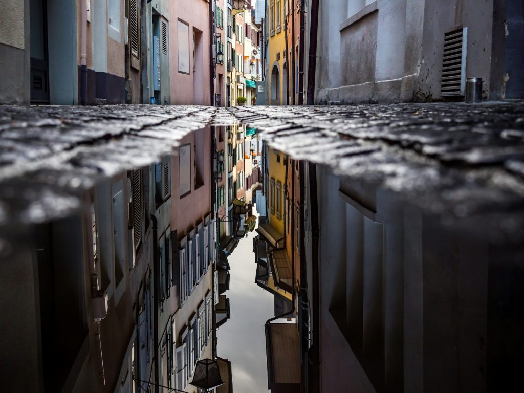 Narrow colorful alley in Morges reflected in a rain puddle on the cobblestone street.