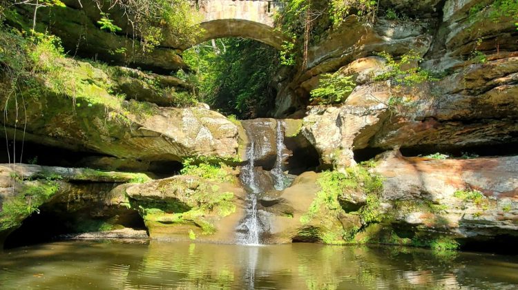 A waterfall flowing beneath a footbridge surrounded by forest at Hocking Hills State Park in Ohio