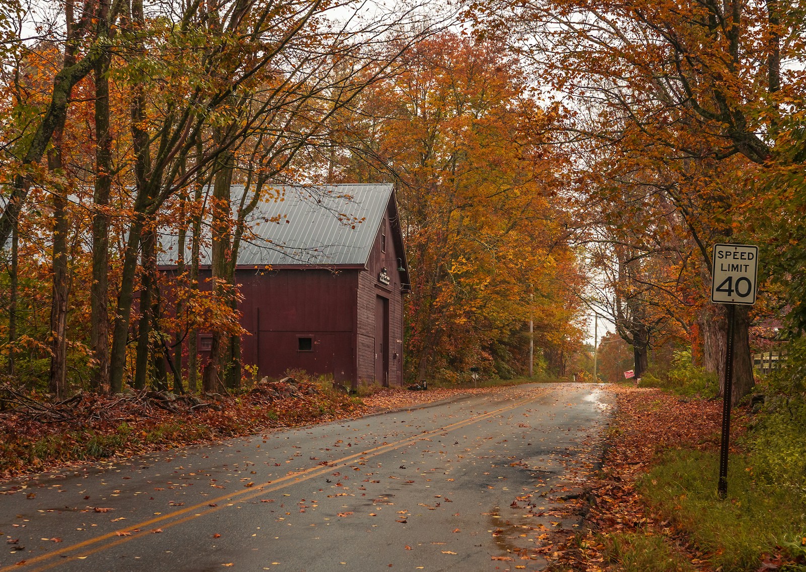 A quiet two-lane country road lined with orange autumn trees, passing a red barn and a speed limit sign in Massachusetts.