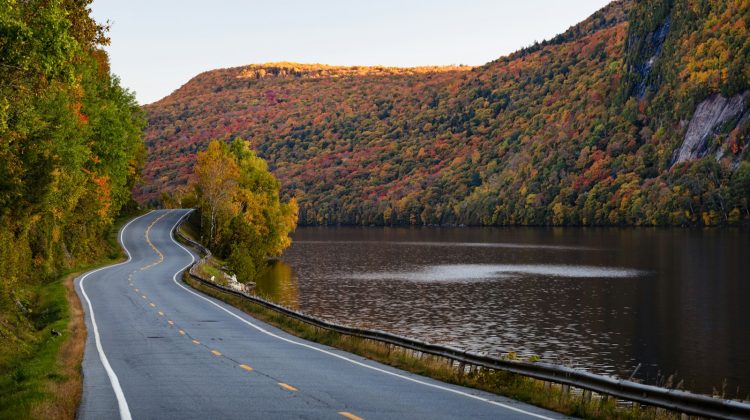 A winding road curves along a lake beneath hillsides of red, orange and yellow autumn foliage in New Hampshire.